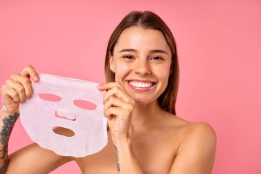 Güzel, mutlu bir kadın pembe arka planda nemlendirici yüz maskesi kullanıyor. Cilt bakımı, kozmetoloji, güzellik konsepti.