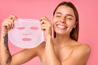 Güzel, mutlu bir kadın pembe arka planda nemlendirici yüz maskesi kullanıyor. Cilt bakımı, kozmetoloji, güzellik konsepti.