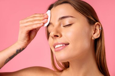 Beyaz tenli beyaz bir kadın pamuk pedlerle yüzünü tonerle siliyor ya da pembe arka planda izole edilmiş makyajı kaldırıyor. Yüz bakımı. Spa tedavileri ve kozmetoloji.