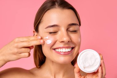 Yüzünde nemlendirici ya da pembe arka planda izole edilmiş güneş kremi kullanan beyaz tenli bir kadın. Yüz bakımı. Spa tedavileri ve kozmetoloji.