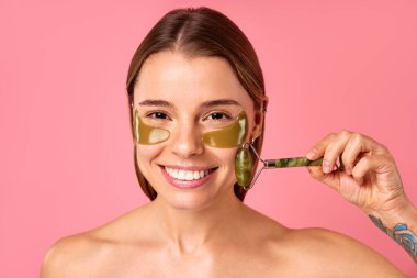 Göz altı nemlendirici bantları olan güzel bir kadın pembe arka planda yeşil bir masözle masaj yapıyor. Yüz bakımı, kozmetoloji, güzellik konsepti..