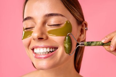 Göz altı nemlendirici bantları olan güzel bir kadın pembe arka planda yeşil bir masözle masaj yapıyor. Yüz bakımı, kozmetoloji, güzellik konsepti..