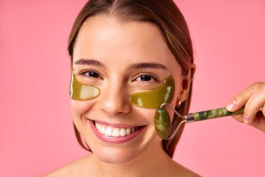 Göz altı nemlendirici bantları olan güzel bir kadın pembe arka planda yeşil bir masözle masaj yapıyor. Yüz bakımı, kozmetoloji, güzellik konsepti..