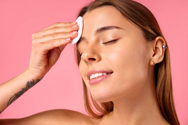 Белая женщина с чистой светящейся кожей с помощью хлопчатобумажных прокладок, вытирая лицо тонером или удаляя макияж изолирован на розовом фоне. Уход за кожей лица. СПА процедуры и косметология.