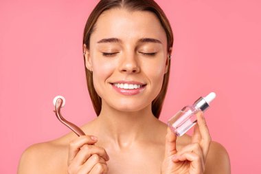 Güzel bir kadın yüz gerdirme için masaj yapıyor ve kırışıklık önleyici olarak mikro terapi ve pembe arka planda izole edilmiş serum kullanıyor. Yüz bakımı, kozmetoloji, güzellik konsepti..