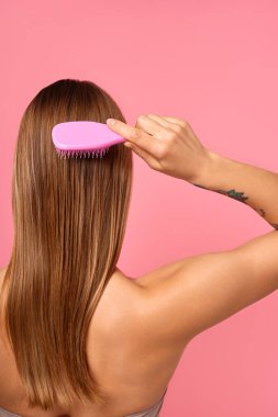 Görüntü, ıslak saçlarını taramak için pembe tarak kullanan bir kadının kolundaki dövmeyi gösteriyor. Günlük rutinlerde saç bakımı, hijyen ve kişisel hizmetin önemini vurgular.