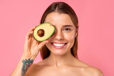 Dövmeli genç bir kadın pembe bir arka planda avokadoyla mutlu bir şekilde poz veriyor, sağlığı, güzelliği, cildini, beslenmeyi ve pozitif bir yaşam tarzını sembolize ediyor. Gülümsemesi ve görünüşü mutluluk saçıyor.