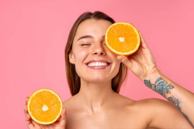 Neşeli bir kadın, parlak bir gülümsemeyle, pembe arka planda iki taze turuncu yarısıyla doğal cilt bakımını ve sağlıklı bir yaşam tarzını teşvik ediyor. Pozitif enerji yayıyor ve refahı savunuyor.