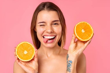 Neşeli bir kadın, parlak bir gülümsemeyle, pembe arka planda iki taze turuncu yarısıyla doğal cilt bakımını ve sağlıklı bir yaşam tarzını teşvik ediyor. Pozitif enerji yayıyor ve refahı savunuyor.