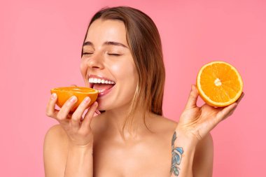 Neşeli bir kadın, parlak bir gülümsemeyle, pembe arka planda iki taze turuncu yarısıyla doğal cilt bakımını ve sağlıklı bir yaşam tarzını teşvik ediyor. Pozitif enerji yayıyor ve refahı savunuyor.