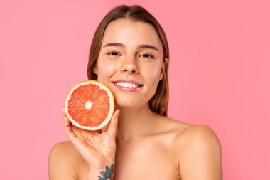 Açık tenli ve doğal makyajlı neşeli bir kadın, pembe arka planda taze bir greyfurt tutarken gülümsüyor. Bu görüntü güzellik ve refah ile ilgili konular için mükemmeldir.