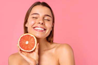 Açık tenli ve doğal makyajlı neşeli bir kadın, pembe arka planda taze bir greyfurt tutarken gülümsüyor. Bu görüntü güzellik ve refah ile ilgili konular için mükemmeldir.
