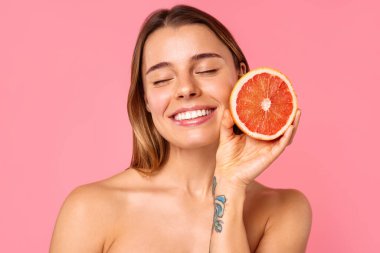 Açık tenli ve doğal makyajlı neşeli bir kadın, pembe arka planda taze bir greyfurt tutarken gülümsüyor. Bu görüntü güzellik ve refah ile ilgili konular için mükemmeldir.
