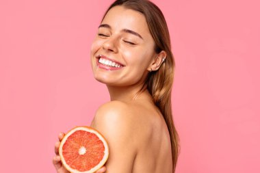 Açık tenli ve doğal makyajlı neşeli bir kadın, pembe arka planda taze bir greyfurt tutarken gülümsüyor. Bu görüntü güzellik ve refah ile ilgili konular için mükemmeldir.