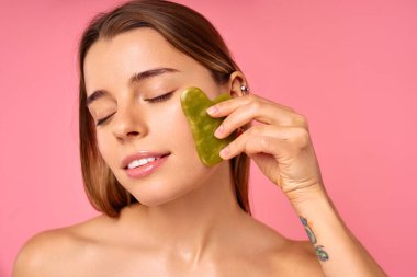 Bir kadın, çenesinde yeşim bir yüz silindiri ile cildini şımartan kişisel bakım ve güzellik rutinine düşkündür. Pembe arka planlı sakin ortam rahatlatıcı bir cilt bakımı için ortam oluşturur