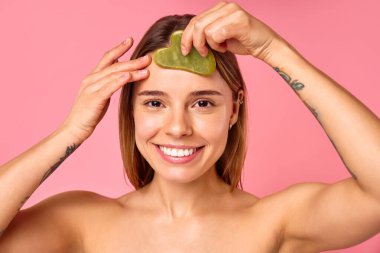 Bir kadın, çenesinde yeşim bir yüz silindiri ile cildini şımartan kişisel bakım ve güzellik rutinine düşkündür. Pembe arka planlı sakin ortam rahatlatıcı bir cilt bakımı için ortam oluşturur