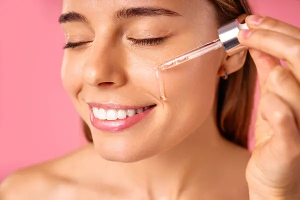 stock image Beautiful woman face, natural clean and fresh glowing skin, applying anti-aging collagen serum. Hands hold serum with active herbal ingredients vitamin C in a dropper, on a pink background.