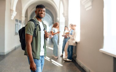 Kalabalık bir üniversite koridorunda, sırt çantalı genç bir erkek öğrenci, arka planda odaklanmış sınıf arkadaşlarıyla tezat kurarak dinamik bir üniversite ortamını yansıtıyor.