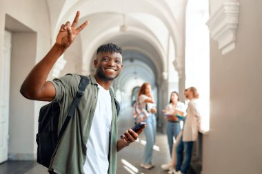 Canlı bir üniversite koridorunda, rahat giyinmiş genç bir adam bir telefon tutar ve barış işareti gösterir. Öðrenci arkadaslarý, akademik canlý bir atmosfer vurguluyor