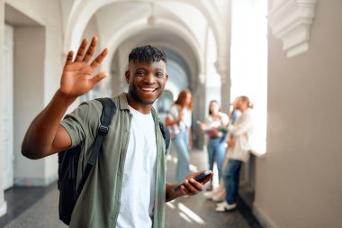 Gülümseyen, el sallayan ve elinde akıllı telefonuyla kampüs koridorunda arkadaşlarıyla sohbet eden bir üniversite öğrencisi. Sahne, yoğun koridorda mutlu bir anı yakalıyor.