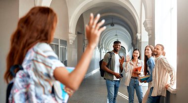 Üniversite koridorunda bir grup genç yetişkin gülümsüyor ve el sallıyor, arkadaşlıklarını ve hayat dolu kampüs yaşamlarını sergiliyor. Etkileşimleri bir topluluk ve beraberlik hissini yansıtıyor.