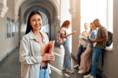Genç bir öğrenci, elinde kitaplarla sohbet eden arkadaşlarıyla çevrili bir üniversite koridoruna güven yayıyor. Sahne, canlı kampüs yaşamının bir anını yakalıyor.