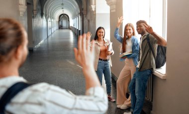 Üniversite koridorunda bir grup genç yetişkin gülümsüyor ve el sallıyor, arkadaşlıklarını ve hayat dolu kampüs yaşamlarını sergiliyor. Etkileşimleri bir topluluk ve beraberlik hissini yansıtıyor.