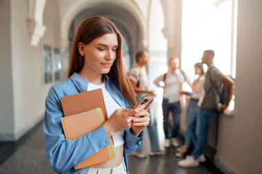 Üniversite koridorunda, bir kız öğrenci, sohbet eden sınıf arkadaşlarıyla çevrili klasörlü ve akıllı telefonlu birden fazla iş yapıyor. Modern eğitim uygulamaları ve akran etkileşimine girerler.