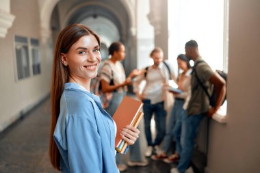 Kendinden emin bir öğrenci meşgul bir üniversite koridorunda, sohbet eden bir grup akran arasında kitap tutarak, üniversite hayatının eğitim, sosyal ve akademik yönlerini sembolize ederek tasvir edilir.