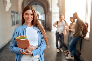 Kendinden emin bir öğrenci meşgul bir üniversite koridorunda, sohbet eden bir grup akran arasında kitap tutarak, üniversite hayatının eğitim, sosyal ve akademik yönlerini sembolize ederek tasvir edilir.