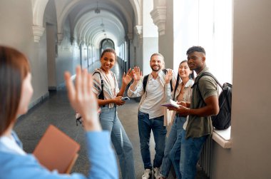 Üniversite koridorunda bir grup genç yetişkin gülümsüyor ve el sallıyor, arkadaşlıklarını ve hayat dolu kampüs yaşamlarını sergiliyor. Etkileşimleri bir topluluk ve beraberlik hissini yansıtıyor.