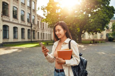 Genç bir kız öğrenci kampüste modern akademik hayattan zevk alıyor, kitap taşırken akıllı telefon kullanıyor, canlı bir gençlik öğrenim ve öğrenim kültürünü sembolize ediyor.