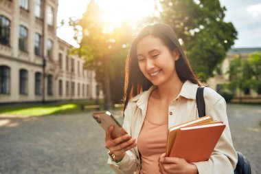 Genç bir kız öğrenci kampüste modern akademik hayattan zevk alıyor, kitap taşırken akıllı telefon kullanıyor, canlı bir gençlik öğrenim ve öğrenim kültürünü sembolize ediyor.