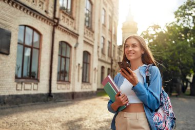 Genç bir kız öğrenci kampüste modern akademik hayattan zevk alıyor, kitap taşırken akıllı telefon kullanıyor, canlı bir gençlik öğrenim ve öğrenim kültürünü sembolize ediyor.