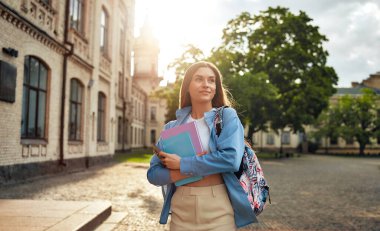 Öğleden sonra üniversite kampüsünde yürürken, elinde kitap ve sırt çantasıyla akademik isteklerini sembolize eden bir kız, umut ve öğrenmeye olan bağlılığını somutlaştırıyor.