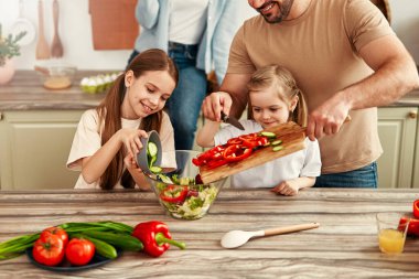 Sıcak bir mutfakta, ebeveynler ve çocuklar taze bir sebze salatası hazırlarken, aile zamanlarını el üstünde tutarken ve sıcak bir atmosferde birlikte yemek pişirirken birbirlerine bağlanırlar.