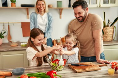 Sıcak bir mutfakta, ebeveynler ve çocuklar taze bir sebze salatası hazırlarken, aile zamanlarını el üstünde tutarken ve sıcak bir atmosferde birlikte yemek pişirirken birbirlerine bağlanırlar.