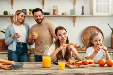 Mutfak masasında taze portakallarla oynayan iki gülümseyen çocuk anne babaları arka planda sıcak bir aile bağı yaratıyor.