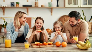 Modern bir mutfakta, kahvaltıda portakal ve meyve suyuyla dört kişilik mutlu bir aile, sevgi ve birliktelik dolu sıcak ve samimi bir atmosfer yaratıyor.