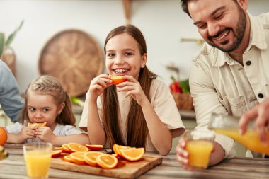 Modern bir mutfakta, kahvaltıda portakal ve meyve suyuyla dört kişilik mutlu bir aile, sevgi ve birliktelik dolu sıcak ve samimi bir atmosfer yaratıyor.