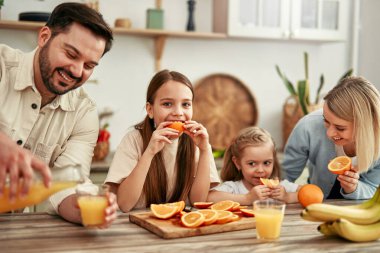 Modern bir mutfakta, kahvaltıda portakal ve meyve suyuyla dört kişilik mutlu bir aile, sevgi ve birliktelik dolu sıcak ve samimi bir atmosfer yaratıyor.