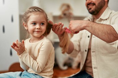 Sıcak bir yatak odasında, bir baba kızının saçlarını nazikçe tarar neşeli bir dekor ve yumuşak bir aydınlatma arasında, sevgi dolu bir anı gösterir. Babalar günün kutlu olsun..