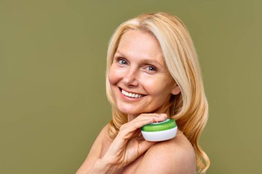 Sarışın, neşeli, orta yaşlı bir kadın yeşil arka planda güzellik ve sağlık için cilt bakım kremi reklamları yapıyor, neşe ve pozitiflik saçıyor. Kozmetik, kaplıca ve cilt bakımı.