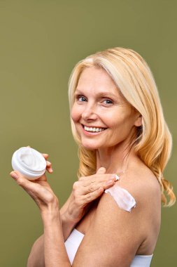 Omzuna nemlendirici krem sürerken gülümseyen olgun bir kadının görüntüsü sağlıklı bir cilt bakımını ve ışıl ışıl cilt için anti-yaşlanma tedavisini somutlaştırır.