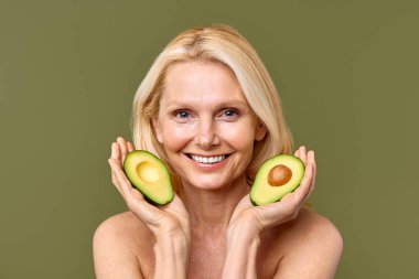 Olgun kadın, elinde avokado yarısıyla zeytin zemininde güven içinde poz veriyor. Kozmetoloji, cilt bakımı ve spa tedavileri. Kaldırma ve estetik ameliyat.