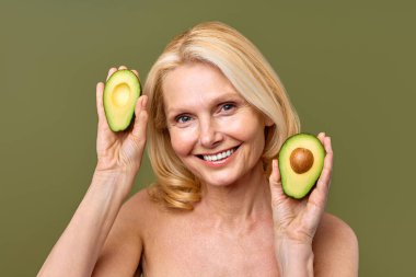 Olgun kadın, elinde avokado yarısıyla zeytin zemininde güven içinde poz veriyor. Kozmetoloji, cilt bakımı ve spa tedavileri. Kaldırma ve estetik ameliyat.