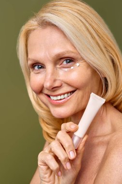 Huzurlu bir ifadeye sahip yaşlı bir kadın gözlerinin etrafına bir tüp cilt bakımı tutarak doğal güzelliği ve özsaygıyı zeytin arka planına karşı teşvik ediyor. Kozmetik, kaplıca ve cilt bakımı.