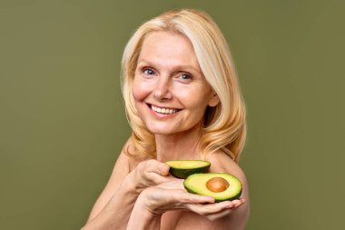 Olgun kadın, elinde avokado yarısıyla zeytin zemininde güven içinde poz veriyor. Kozmetoloji, cilt bakımı ve spa tedavileri. Kaldırma ve estetik ameliyat.