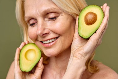 Olgun kadın, elinde avokado yarısıyla zeytin zemininde güven içinde poz veriyor. Kozmetoloji, cilt bakımı ve spa tedavileri. Kaldırma ve estetik ameliyat.