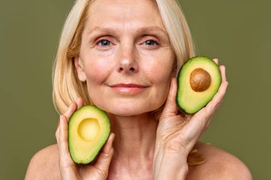 Olgun kadın, elinde avokado yarısıyla zeytin zemininde güven içinde poz veriyor. Kozmetoloji, cilt bakımı ve spa tedavileri. Kaldırma ve estetik ameliyat.